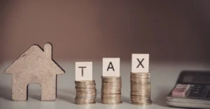 Stacks of coins labeled tax next to a house model and calculator representing IRS property seizure for unpaid taxes.