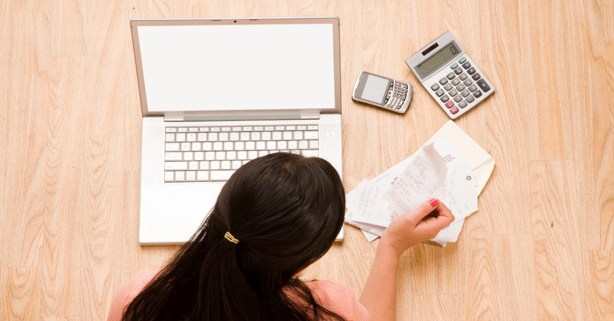 Woman sitting on the floor with a laptop, receipts, and calculators, organizing financial records.
