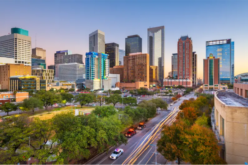 Downtown Houston skyline at dusk with modern skyscrapers, busy streets, and trees in the foreground.